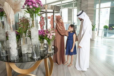 Parents' follow-up and care for their children, spending happy times with family, gestures of happiness and joy, an Emirati Gulf Arab mother wearing an abaya and hijab stands with her husband at the hotel reception entrance, placing a flower in her daughter's hair, preparing to enjoy a pleasant family trip.