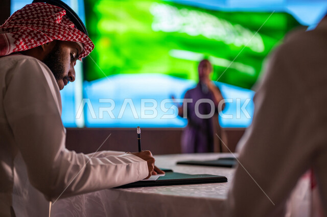 سيدة عربية خليجية سعودية تقف امام خلفية لعلم المملكة العربية السعودية ، القاء كلمة ترحيبية في مؤتمر سعودي ، الاحتفال باليوم الوطني السعودي ، رجل سعودي خليجي يدون الملاحظات ، تقييم الموهوبين السعوديين ،  مؤتمرات و ندوات في السعودية
