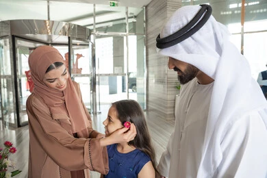 Parents' care for their children and their upbringing, spending happy times with family, gestures of happiness and joy, an Emirati Gulf Arab mother wearing an abaya and hijab stands with her daughter and husband at the hotel reception entrance, placing a flower in her hair, preparing to enjoy a pleasant family trip.