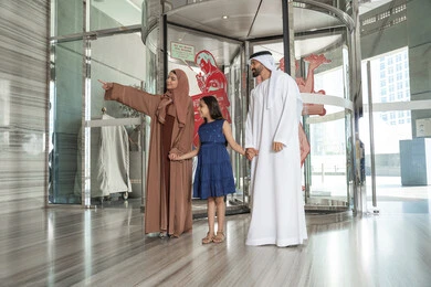 رحلة عائلية ترفيهية، رجل عربي خليجي اماراتي يرتدي الكندورة و الدشداشة يقف برفقه زوجته و ابنته بايماءات السعادة و السرور ، الاشارة الى شيء ما، لحظات دخول عائلة عربية خليجية اماراتية إلى الفندق السياحي