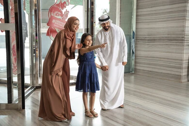 استمتاع الأسرة بوقت مميز، رجل عربي خليجي اماراتي يرتدي الكندورة و الدشداشة يقف برفقه زوجته و ابنته بايماءات السعادة و السرور ، الاشارة الى شيء ما، لحظات دخول عائلة عربية خليجية اماراتية إلى الفندق السياحي