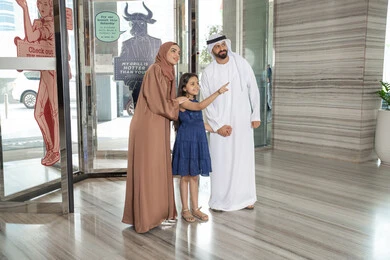 The concept of tourism and travel, an Arab Gulf Emirati man wearing a kandura and a dishdasha stands with his wife and daughter with gestures of happiness and joy, pointing to something, enjoyable family times, moments of an Arab Gulf Emirati family entering a tourist hotel.