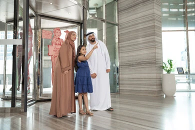 رجل عربي خليجي اماراتي يرتدي الكندورة و الدشداشة يقف برفقه زوجته و ابنته بايماءات السعادة و السرور ، الاشارة الى شيء ما،  أوقات عائلية ممتعة، لحظات دخول عائلة عربية خليجية اماراتية إلى الفندق السياحي ، مفهوم السياحة و السفر