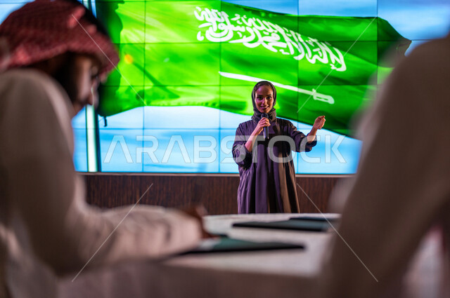 سيدة عربية خليجية سعودية تقف امام خلفية لعلم المملكة العربية السعودية ، القاء كلمة ترحيبية في مؤتمر سعودي ، الاحتفال باليوم الوطني السعودي ، رجل سعودي خليجي يدون الملاحظات ، تقييم الموهوبين السعوديين ،  مؤتمرات و ندوات في السعودية