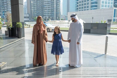 زوجان عربيان خليجيين اماراتيين يرتديان الزي التقليدي يسيران امام الفندق برفقة ابنتهم، قضاء رحلة صيفية ممتعة ومسلية، قضاء وقت ممتع، مفهوم السياحة في إجازة الصيف، الاستعداد للسفر في جولة سياحية