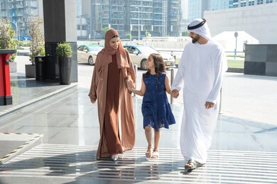 Spending an enjoyable and entertaining summer trip, having a good time, the concept of tourism during the summer vacation, an Emirati Arab couple wearing traditional attire walking in front of the hotel with their daughter, preparing to travel on a sightseeing tour.