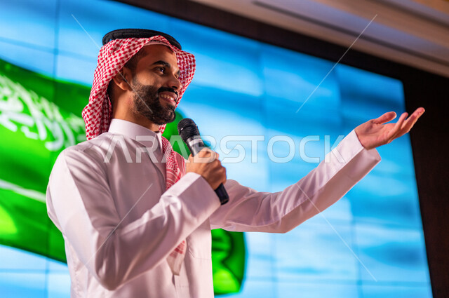 A Saudi Arabian Gulf man standing in front of a background of the flag of the Kingdom of Saudi Arabia, giving a speech at a conference on the Saudi National Day, the ceremony of Saudi talents, the celebration of Saudi Independence Day, conferences and sem