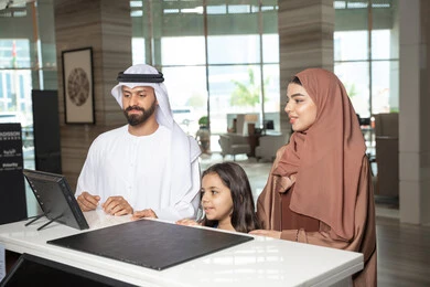 إنهاء إجراءات الحجز داخل المنتجع، الاستمتاع بالعطلة الصيفية، رجل اماراتي يرتدي الكندورة و الغتره يقف برفقة زوجته و ابنته عند مكتب الاستقبال فى احد الفنادق الفاخره، تعابير السعادة و السرور