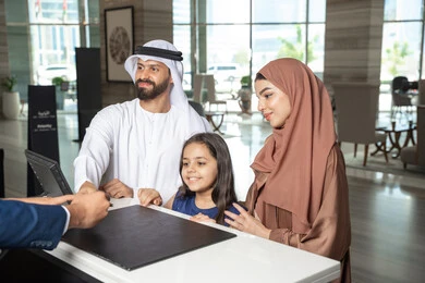 Checking the reservation at the resort, enjoying the summer vacation, an Emirati man wearing the kandura and ghutrah stands with his wife and daughter at the reception desk of a luxury hotel, expressions of happiness and joy.