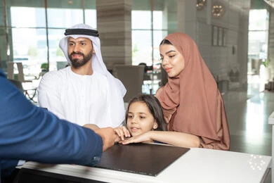 إتمام عملية الحجز في المنتجع، الاستمتاع بالعطلة الصيفية، رجل اماراتي يرتدي الكندورة و الغتره يقف برفقة زوجته و ابنته عند مكتب الاستقبال فى احد الفنادق الفاخره، تعابير السعادة و السرور