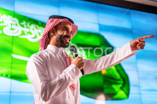 رجل عربي خليجي سعودي يقف امام خلفية لعلم المملكة العربية السعودية ، يشير باصبع السبابة لشيء ما ، حفل الموهوبين السعوديين، مؤتمر عن اليوم الوطني السعودي ، الاحتفال بعيد الاستقلال السعودي ، ذكرى تاسيس المملكة العربية السعودية