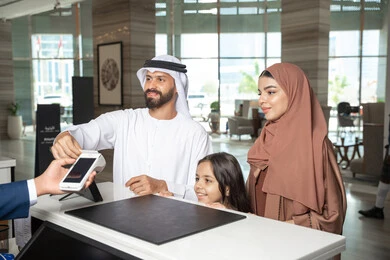 تسديد المدفوعات من خلال بطاقة الصراف، تعابير السعادة و السرور، عطلة نهاية الأسبوع، رجل اماراتي يرتدي الكندورة و الغتره يقف برفقة زوجته و ابنته عند مكتب الاستقبال فى احد الفنادق الفاخره يقوم بتأكيد حجزه داخل المنتجع