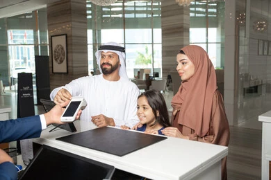 Paying bills using an ATM card, expressions of happiness and joy, the weekend, an Emirati man wearing a kandura and ghutrah standing with his wife and daughter at the reception desk of a luxury hotel confirming his reservation at the resort.