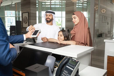 The possibility of paying bills via a debit card, expressions of happiness and joy, the weekend, an Emirati man wearing a kandura and ghutrah stands with his wife and daughter at the reception desk of a luxury hotel confirming his reservation at the resort.