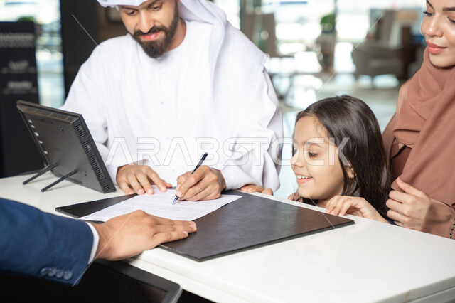 Weekend, a close-up shot of an Emirati man wearing a kandura and ghutrah standing with his wife and daughter at the reception desk of a luxury hotel confirming his reservation at the resort, expressions of happiness and joy.