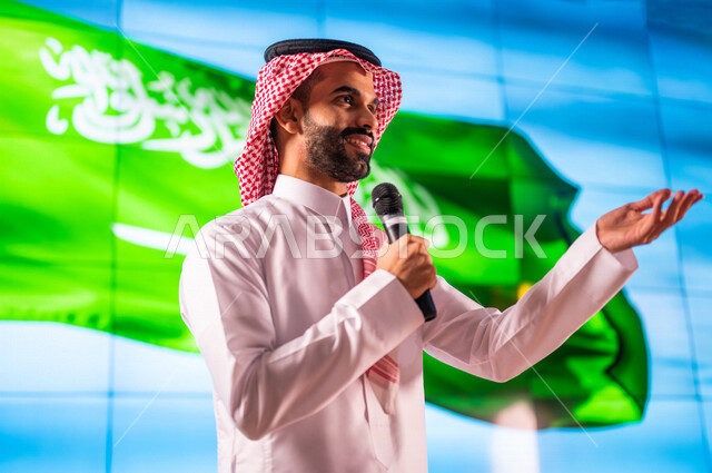 رجل عربي خليجي سعودي يقف امام خلفية لعلم المملكة العربية السعودية ،يقوم بالقاء كلمة في مؤتمر عن اليوم الوطني السعودي ، حفل الموهوبين السعوديين ، الاحتفال بعيد الاستقلال السعودي ، مؤتمرات و  ندوات داخل المملكة العربية السعودية