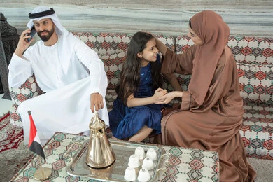 Feelings of safety and love with family, using modern technology, an Arab Gulf Emirati man wearing a kandura and ghutrah sitting in a traditional floor majlis with his daughter and wife, making a phone call via a mobile phone, the concept of caring for and paying attention to children.
