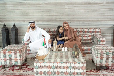 الاشارة الى شيء ما ، المحافظة على الترابط الأسري، قضاء وقت ممتع فى الخارج ، رجل عربي خليجي اماراتي يرتدي الكندورة و الغتره يجلس فى مجلس شعبي ارضي برفقة زوجته و ابنته يقوم باجراء مكالمة هاتفية عبر الجوال ، تعابير السعادة و السرور