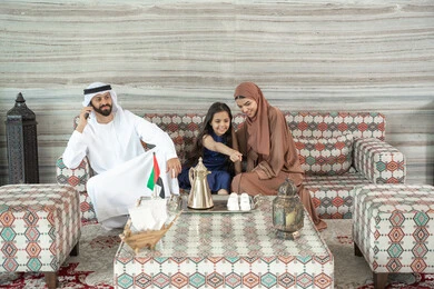 Maintaining family ties, spending enjoyable time outdoors, pointing to something, an Arab Gulf Emirati man wearing a kandura and ghutrah sitting in a traditional ground gathering with his wife and daughter making a phone call via mobile, expressions of happiness and joy.
