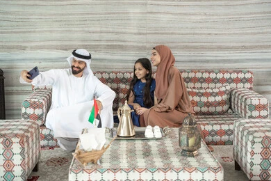قضاء وقت ممتع في الخارج، الترابط الأسري والحفاظ على الاستقرار العائلي، رجل عربي خليجي اماراتي يرتدي الكندورة و الغتره يجلس فى مجلس ارضي شعبي برفقة  زوجته و ابنته  يقوم بالتقاط صور السيلفي عبر الهاتف المحمول ، تعابير السعادة و السرور