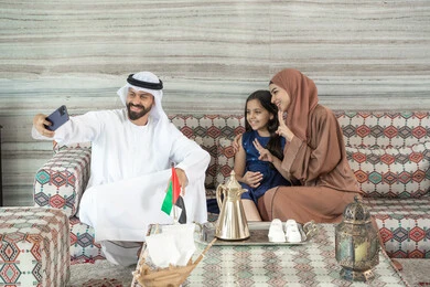 الترابط الأسري والحفاظ على الاستقرار العائلي،مفهوم القبول والموافقه، رجل عربي خليجي اماراتي يرتدي الكندورة و الغتره يجلس فى مجلس ارضي شعبي برفقة  زوجته و ابنته  يقوم بالتقاط صور السيلفي عبر الهاتف المحمول ، تعابير السعادة و السرور