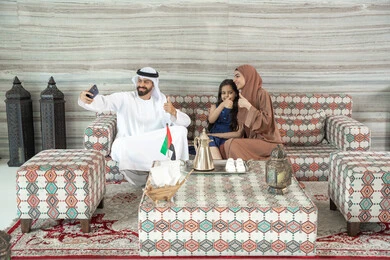 أنشطة ترفيهية مع الأسرة، إشارات بالموافقة والإيجاب، مفهوم الأسرة السعيدة، رجل عربي خليجي اماراتي يرتدي الكندورة و الغتره يجلس فى مجلس ارضي شعبي برفقة  زوجته و ابنته  يقوم بالتقاط صور السيلفي عبر الهاتف المحمول ، تعابير السعادة و السرور