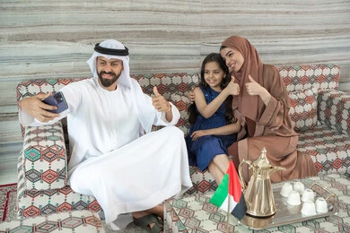 إشارات توحي بالموافقة، أنشطة ترفيهية مع الأسرة، مفهوم الأسرة السعيدة، رجل عربي خليجي اماراتي يرتدي الكندورة و الغتره يجلس فى مجلس ارضي شعبي برفقة  زوجته و ابنته  يقوم بالتقاط صور السيلفي عبر الهاتف المحمول ، تعابير السعادة و السرور