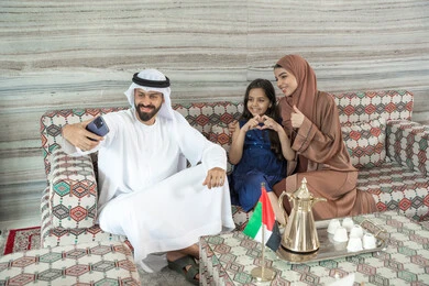 Gestures of approval and acceptance, shaping the hands in the form of a heart, recreational activities with the family, the concept of a happy family, an Emirati Gulf Arab man wearing a kandura and ghutrah sitting in a traditional floor seating area with his wife and daughter, taking selfies with a mobile phone, expressions of happiness and joy.