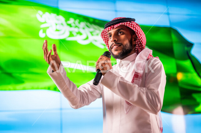 رجل عربي خليجي سعودي يقف امام خلفية لعلم المملكة العربية السعودية ،يقوم بالقاء كلمة في مؤتمر عن اليوم الوطني السعودي ، حفل الموهوبين السعوديين ، الاحتفال بعيد الاستقلال السعودي ، مؤتمرات و  ندوات داخل المملكة العربية السعودية