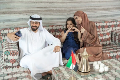 ايماءات الموافقة و القبول، تشكيل اليدين على هيئة قلب، أنشطة ترفيهية مع الأسرة، مفهوم الأسرة السعيدة، رجل عربي خليجي اماراتي يرتدي الكندورة و الغتره يجلس فى مجلس ارضي شعبي برفقة  زوجته و ابنته  يقوم بالتقاط صور السيلفي عبر الهاتف المحمول ، تعابير السعادة و السرور