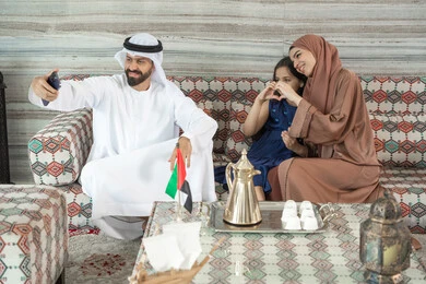 Shaping the hands in the form of a heart, recreational activities with the family, the concept of a happy family, an Emirati Gulf Arab man wearing a kandura and ghutrah sitting in a traditional floor seating area with his wife and daughter, taking selfies with a mobile phone, expressions of happiness and joy. Shaping the hands in the form of a heart, recreational activities with the family, the concept of a happy family, an Emirati Gulf Arab man wearing a kandura and ghutrah sitting in a traditional floor seating area with his wife and daughter, taking selfies with a mobile phone, expressions of happiness and joy.