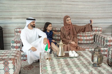 ايماءات يدين على شكل قلب، توثيق اللحظات السعيدة ، أنشطة ترفيهية مع الأسرة، مفهوم الأسرة السعيدة، امراه عربيه خليجيه اماراتية ترتدي العباءة و الحجاب تجلس فى مجلس ارضي شعبي برفقة زوجها و ابنتها تقوم بالتقاط صور السيلفي عبر الهاتف المحمول