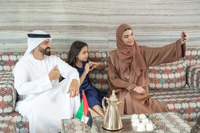 Documenting happy moments, recreational activities with family, the concept of a happy family, an Emirati Gulf Arab woman wearing an abaya and hijab sitting in a traditional floor seating area with her husband and daughter taking selfies with a mobile phone, hand gestures in the shape of a heart.