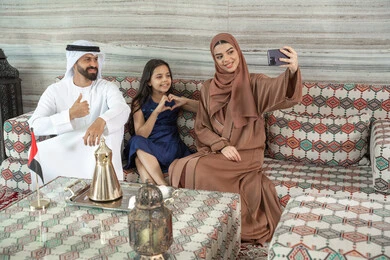 أنشطة ترفيهية مع الأسرة، مفهوم الأسرة السعيدة، امراه عربيه خليجيه اماراتية ترتدي العباءة و الحجاب تجلس فى مجلس ارضي شعبي برفقة زوجها و ابنتها تقوم بالتقاط صور السيلفي عبر الهاتف المحمول ، توثيق اللحظات السعيدة ، ايماءات يدين على شكل قلب