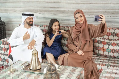 The concept of a happy family, recreational activities with the family, an Arab Gulf Emirati woman wearing an abaya and hijab sitting in a traditional floor seating area with her husband and daughter taking selfies with a mobile phone, documenting happy moments, hand gestures in the shape of a heart.