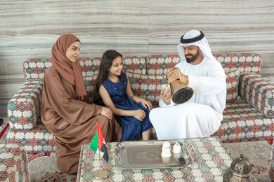 Enjoying outdoor activities, sipping hot drinks, a happy family atmosphere filled with love and affection, spending free time entertaining with the family, an Emirati Gulf Arab man wearing a kandura and ghutrah sitting in a traditional floor seating area with his daughter and wife pouring coffee, gestures of happiness and joy. Enjoying outdoor activities, sipping hot drinks, a happy family atmosphere filled with love and affection, spending free time entertaining with the family, an Emirati Gulf Arab man wearing a kandura and ghutrah sitting in a traditional floor seating area with his daughter and wife pouring coffee, gestures of happiness and joy.