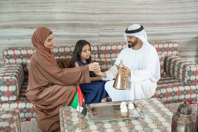 Spending a beautiful time outdoors, sipping hot drinks, a happy family atmosphere filled with love and affection, utilizing free time for entertainment with the family, an Arab Gulf Emirati man wearing a kandura and ghutrah sitting in a traditional floor seating area with his daughter and wife pouring coffee, gestures of happiness and joy.
