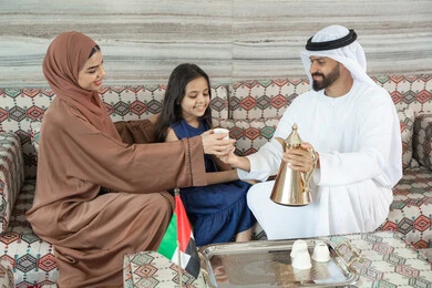 Drinking hot beverages, happy family atmosphere filled with love and affection, spending enjoyable times outdoors, utilizing free time for entertainment with the family, an Emirati Gulf Arab man wearing a kandura and ghutrah sitting in a traditional floor seating area with his daughter and wife pouring coffee, gestures of happiness and joy.