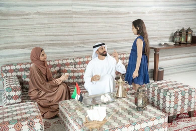 أجواء عائلية سعيدة مليئة بالحب والحنان، قضاء أوقات ممتعة في الخارج، استغلال وقت الفراغ في التسلية مع الاسرة، الاستمتاع بتناول المشروبات الساخنة، رجل عربي خليجي اماراتي يرتدي الكندورة و الغتره يجلس فى مجلس ارضي شعبي برفقة ابنته و زوجته، ايماءات السعادة و السرور