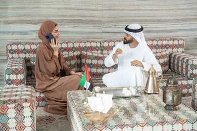 الدردشة والتواصل مع الاهل والأصدقاء، قضاء أوقات ممتعة، تناول وشرب المشروبات الساخنة، امراه عربيه خليجيه اماراتية ترتدي العباءة و الحجاب تجلس فى مجلس ارضي شعبي برفقة زوجها تقوم باجراء مكالمه هاتفيه عبر الهاتف المحمول، ايماءات السعادة و السرور