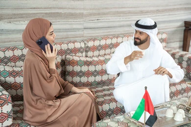 Spending enjoyable times, having and drinking hot beverages, an Emirati Gulf Arab woman wearing an abaya and hijab sits in a traditional floor seating area with her husband, making a phone call on her mobile phone, with gestures of happiness and joy.