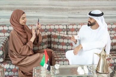 Gestures of happiness and joy, using modern technology and techniques, documenting happy moments by taking commemorative photos, an Arab Gulf Emirati woman wearing an abaya and hijab sitting in a traditional floor seating area photographing her husband using a mobile phone. Gestures of happiness and joy, using modern technology and techniques, documenting happy moments by taking commemorative photos, an Arab Gulf Emirati woman wearing an abaya and hijab sitting in a traditional floor seating area photographing her husband using a mobile phone.
