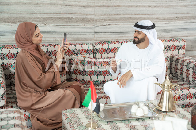 استخدام اتكنولوجيا والتقنيات الحديثة، توثيق اللحظات السعيدة من خلال التقاط الصور التذكارية، امراه عربيه خليجيه اماراتية ترتدي العباءة و الحجاب  تجلس فى مجلس ارضي شعبي تقوم بتصوير زوجها باستخدام الهاتف المحمول ، ايماءات السعاده و السرور