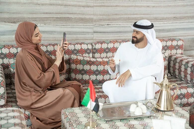 استخدام اتكنولوجيا والتقنيات الحديثة، توثيق اللحظات السعيدة من خلال التقاط الصور التذكارية، امراه عربيه خليجيه اماراتية ترتدي العباءة و الحجاب  تجلس فى مجلس ارضي شعبي تقوم بتصوير زوجها باستخدام الهاتف المحمول ، ايماءات السعاده و السرور