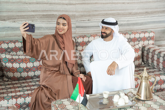توثيق اللحظات العائلية السعيدة، إجراء مكالمة فيديو أونلاين، مفهوم الأمان والإستقرار العائلي، زوجان عربيان خليجيان اماراتيان يرتديان الزي التقليدي يجلسان فى مجلس ارضي شعبي يلتقطان صورة تذكارية من خلال الهاتف المحمول، المحافظة على الترابط الأسري