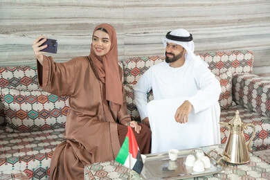 توثيق اللحظات العائلية السعيدة، إجراء مكالمة فيديو أونلاين، مفهوم الأمان والإستقرار العائلي، زوجان عربيان خليجيان اماراتيان يرتديان الزي التقليدي يجلسان فى مجلس ارضي شعبي يلتقطان صورة تذكارية من خلال الهاتف المحمول، المحافظة على الترابط الأسري