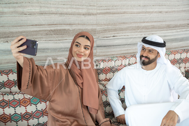 إجراء مكالمة فيديو أونلاين، مفهوم الأمان والإستقرار العائلي، توثيق اللحظات العائلية السعيدة،زوجان عربيان خليجيان اماراتيان يرتديان الزي التقليدي يجلسان فى مجلس ارضي شعبي يلتقطان صورة تذكارية من خلال الهاتف المحمول، المحافظة على الترابط الأسري