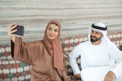 إجراء مكالمة فيديو أونلاين، مفهوم الأمان والإستقرار العائلي، توثيق اللحظات العائلية السعيدة،زوجان عربيان خليجيان اماراتيان يرتديان الزي التقليدي يجلسان فى مجلس ارضي شعبي يلتقطان صورة تذكارية من خلال الهاتف المحمول، المحافظة على الترابط الأسري