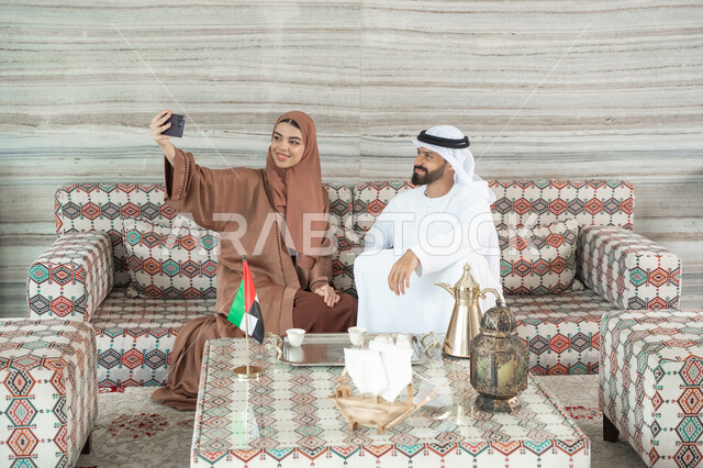 The concept of family security and stability, making an online video call, documenting happy family moments, an Emirati Arab couple wearing traditional attire sitting in a popular floor seating area taking a commemorative photo through a mobile phone, maintaining family cohesion.