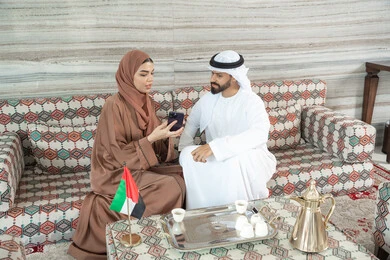 تقوية العلاقات الزوجية وروابط الحب والمودة، زوجان عربيان خليجيان اماراتيان يرتديان الزي التقليدي يجلسان فى مجلس ارضي شعبي يستمتعان باستخدام الهاتف المحمول، متابعة أخبار مواقع التواصل الاجتماعي، مشاهدة الافلام والمسلسلات عبر الجوال